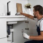 Plombier de Dépan’Services Gaz & Eau intervenant sur le siphon et la tuyauterie sous un évier de cuisine moderne.